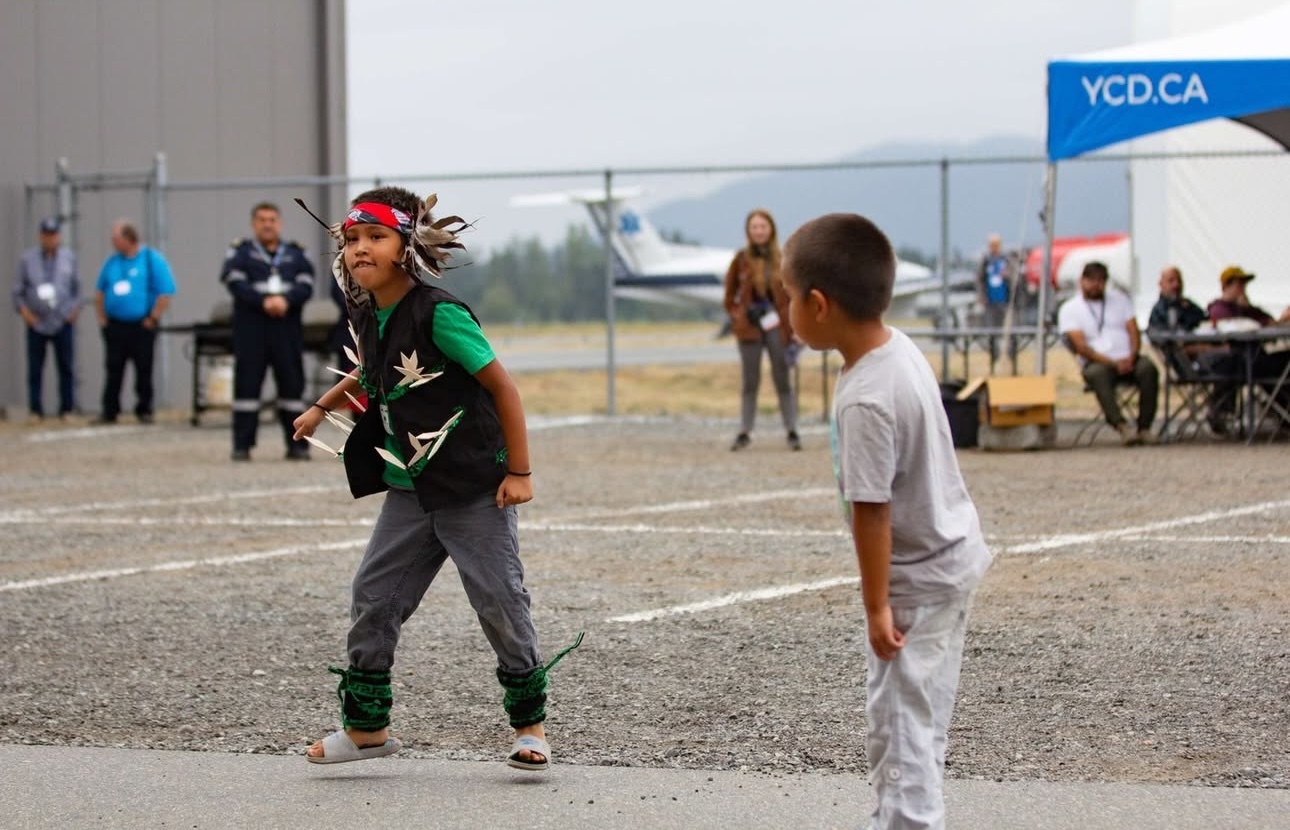 Deux enfants dansent sur le tarmac d’un aéroport, tandis qu’un groupe de personnes les regarde. 