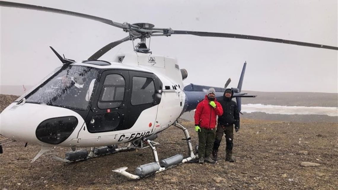 Deux chercheurs vêtus de vêtements adaptés au froid, debout devant un hélicoptère sur un paysage rocheux plat. 