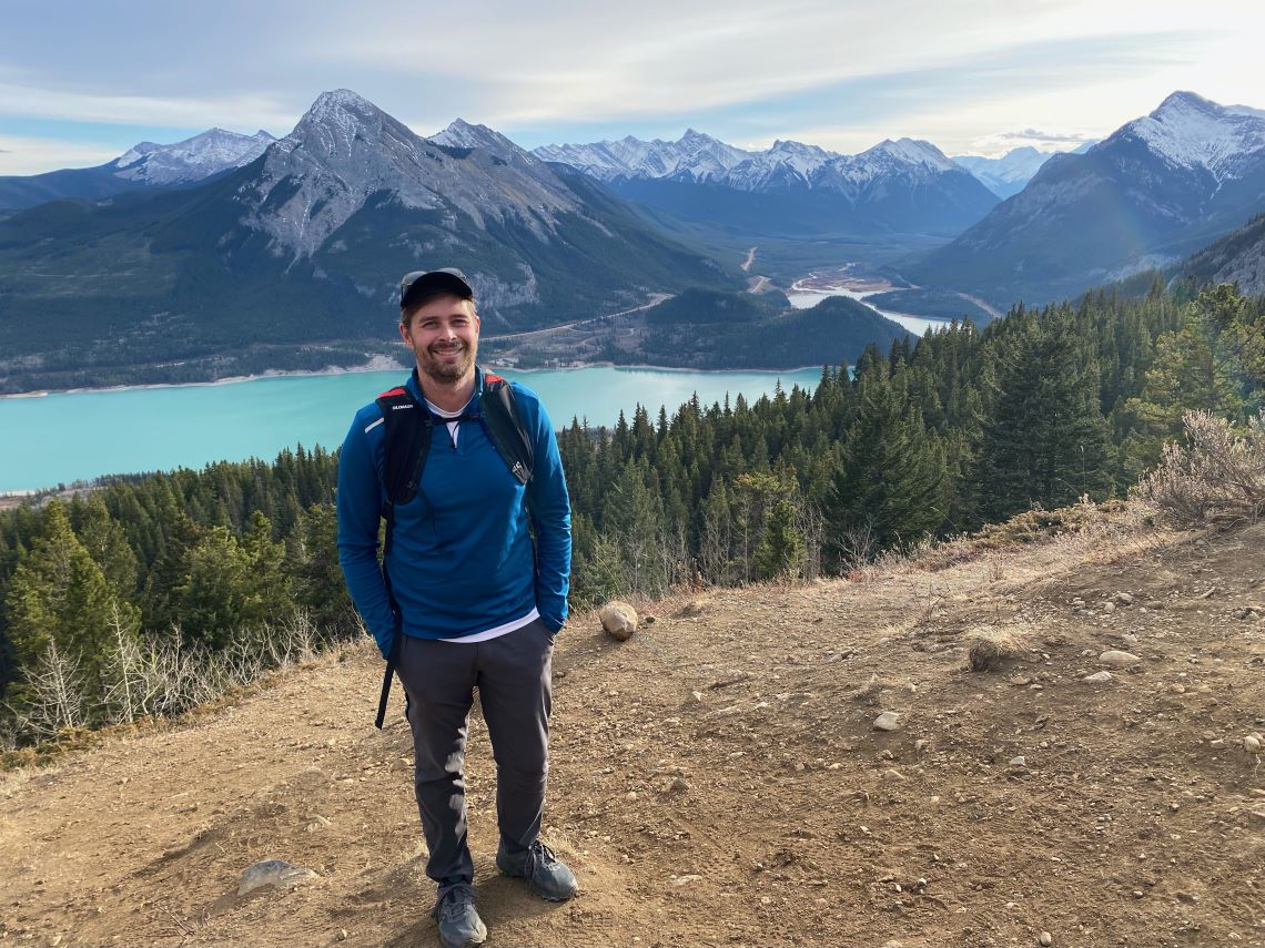 Un homme debout sur une pente, avec une forêt, une rivière et une chaîne de montagnes en arrière-plan.