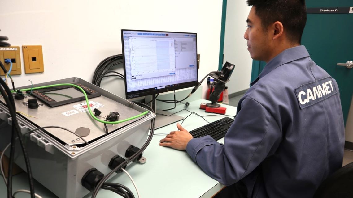 Assis à un poste de travail dans un laboratoire, un scientifique examine des données à l’écran d’un ordinateur branché à du matériel électronique.