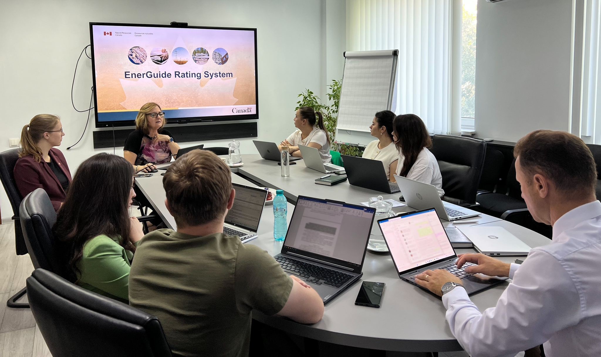 Une réunion d’affaires avec huit personnes assises autour d’une table de conférence, se concentrant sur une présentation affichée sur un grand écran.