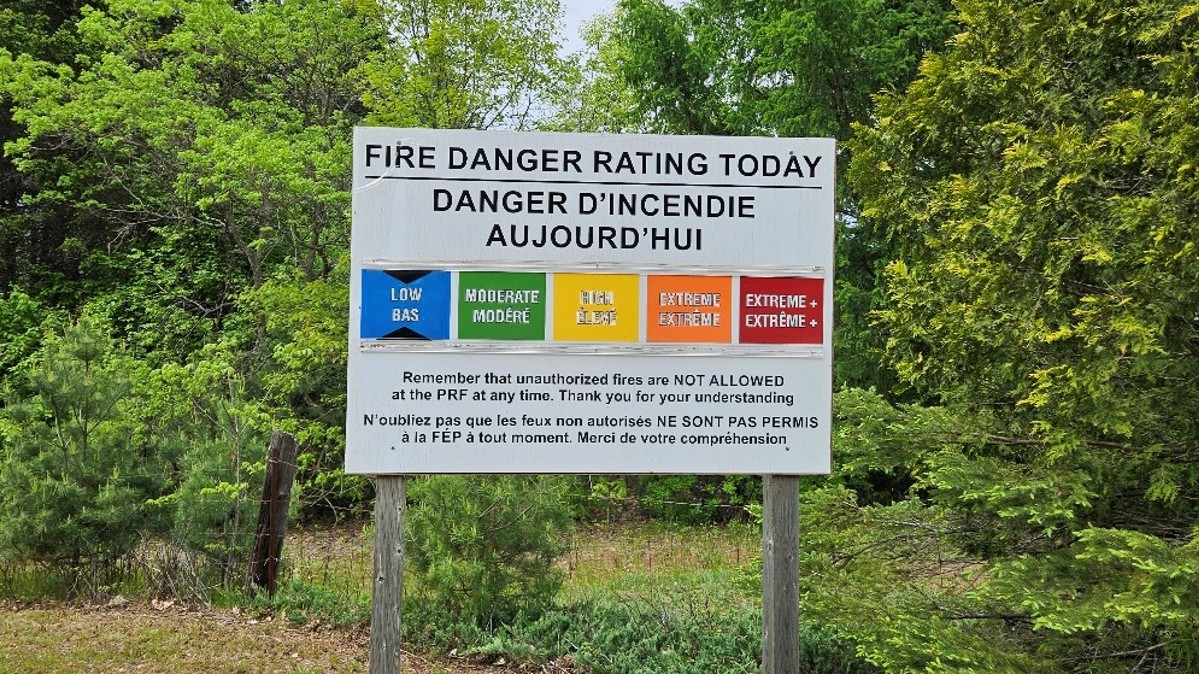 Un panneau bilingue dans un secteur boisé indique le danger d’incendie selon une échelle de cinq couleurs allant de bas à extrême plus.