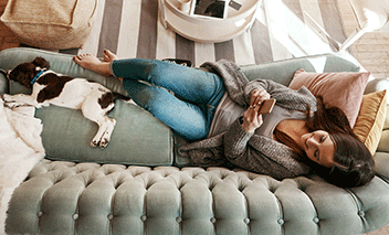Une femme qui se détend sur un canapé à la maison, en utilisant son téléphone, accompagnée d’un chien.