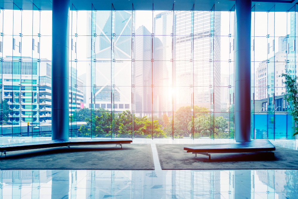 Sur cette image on voit une vue de l'intérieur d'un bâtiment moderne, regardant à travers un grand mur de verre avec le soleil qui brille à l’extérieur. D'autres bâtiments peuvent être vus au loin.