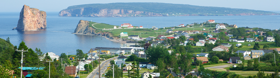 Vue du village côtier de Percé, au Québec, avec une formation rocheuse abrupte à gauche (Rocher Percé) et l'île Bonaventure en arrière-plan.