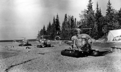 Trois hélicoptères sur le sol avec une tente à la gauche et des arbres dans l’arrière-plan.