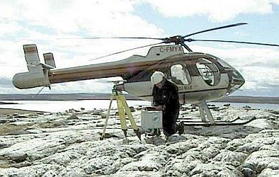 Technicien å genou sur un affleurement rocheux, devant un hélicoptère, qui prend des mesures avec un gravimètre CG-5