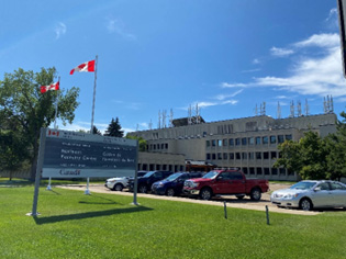 Un immeuble en béton et un ciel bleu vif en arrière-plan. Devant l’immeuble sont deux drapeaux canadiens qui flottent au vent, des voitures dans un parc de stationnement et une enseigne qui porte les mots « Northern Forestry Centre / Centre de foresterie du Nord ». 