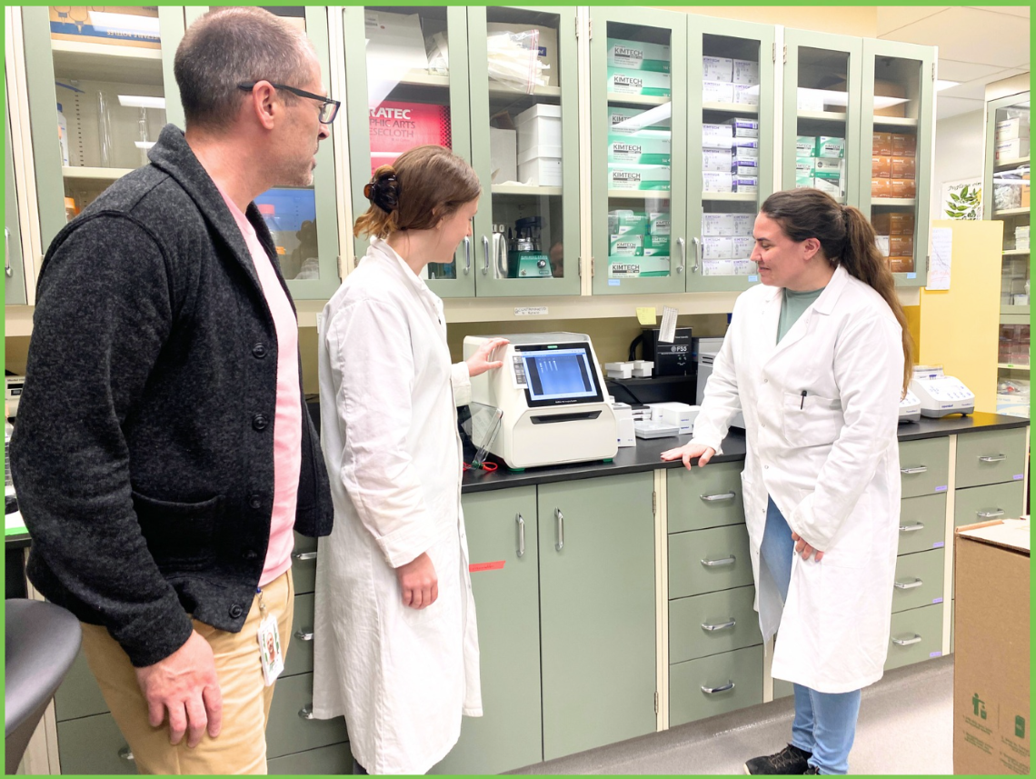 Homme en pull-over et deux femmes en blouse de laboratoire regardent une machine d’analyse d’ADN.