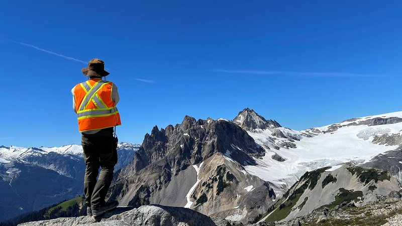 Un chercheur portant une veste de sécurité tourne le dos à la caméra tandis qu’il observe le sommet enneigé du mont Vista.